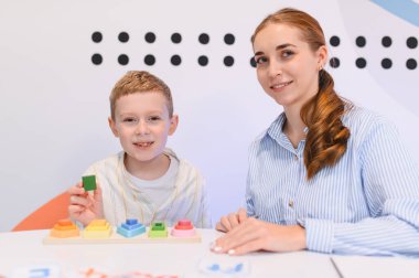 Genç bir çocuk renkli eğitim oyuncaklarıyla oynarken ilgili bir öğretmen ona yardım ediyor, gelişimi teşvik ediyor ve terapi görüyor.