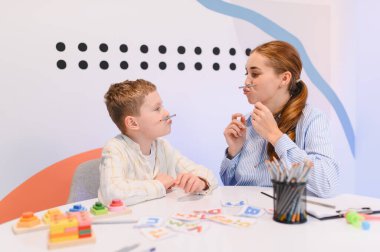 Genç bir çocuk ve terapist konuşma gelişimini ve konuşma becerilerini geliştirmek için kalemlerle eklem egzersizleri yapıyor, klinikte öğrenim görüyor.