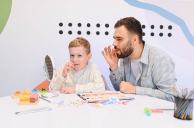 Konuşma terapistiyle konuşma egzersizi yapan bir çocuk. Parlak bir odada iletişim becerilerinin geliştirilmesi ve gelişim ihtiyaçlarının giderilmesi