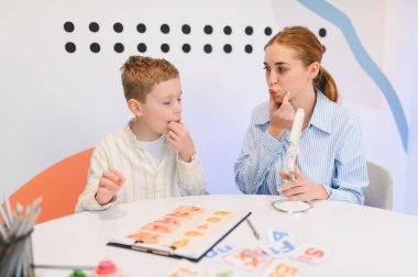 Konuşma terapisti terapi sırasında konuşma egzersizleri yapan bir çocuğa yardım ediyor. Pratik için ağız şekli kartları ve ayna kullanıyorlar.