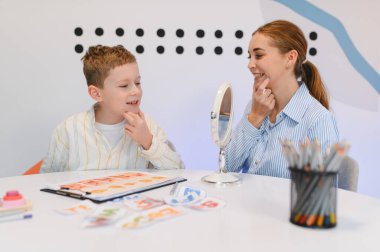 Genç bir çocuk, ayna kullanarak kadın bir logopediciyle yaptığı konuşma terapisi sırasında telaffuz ve konuşma becerileri öğreniyor.