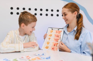 Genç bir konuşma terapisti gülümseyen bir çocuğa çeşitli ağız pozisyonları göstererek konuşma egzersizleri öğretiyor.