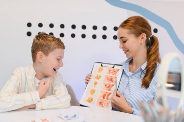 Konuşma terapisti, gelişim oturumunda bir grafikteki yüz ifadelerini ve ağız hareketlerini açıklarken gülümseyen bir çocuk.