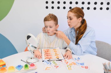 Genç bir çocukla çalışan konuşma terapisti, modern bir klinikte ayna ve eğitim materyalleri kullanarak konuşma sanatında ona yardımcı oluyor.