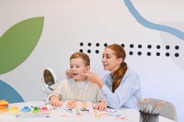 Konuşma terapisti genç bir çocukla çalışıyor. Ders sırasında ayna ve eğitim kartları kullanarak sesleri dile getirmeye yardım ediyor.