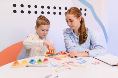 Genç çocuk renkli eğitim oyuncaklarıyla oynarken, bayan eğitmen gelişimsel görevlerde öğretiyor ve yardımcı oluyor.