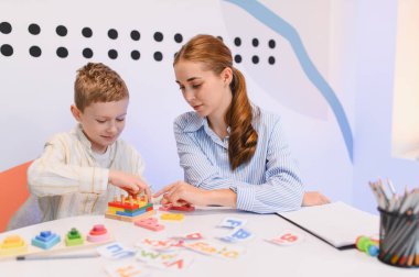 Eğitimsel destek alan, renkli ahşap bulmacalarla uğraşan ve parlak bir odadaki bir masada öğrenme materyalleri kullanan küçük bir çocuk.