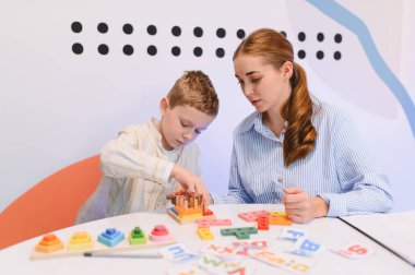 Genç bir çocuk ahşap eğitici oyuncaklarla oynarken, bayan bir öğretmen ona parlak bir sınıf ortamında yardımcı oluyor, erken gelişimi teşvik ediyor.