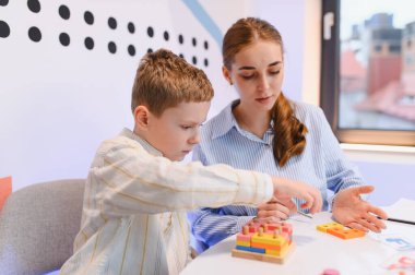 Renkli tahta sıralama taşlarıyla oynayan genç bir çocuk, konsantrasyon ve motor becerileri geliştirmede yardımcı olan bir bayan öğretmen.