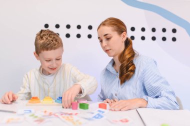 Mandal tahtasında renkli tahta blokları istiflemeye odaklanan bir çocuk, kadın bir öğretmenle el göz koordinasyonunu geliştiriyor.