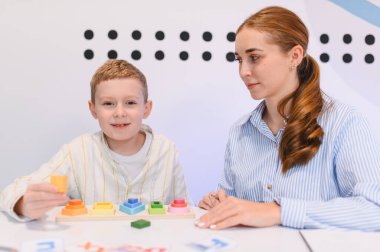 Genç erkek çocuk, renkli ahşap geometrik blokları kadın akıl hocasıyla sıralarken gülümsüyor ve erken eğitime odaklanıyor.