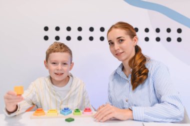 Çocuk renkli ahşap bloklarla oynuyor, sıralama ve istifleme pratiği yapıyor. Sınıfta çocuk gelişimine yardımcı olan öğretmen