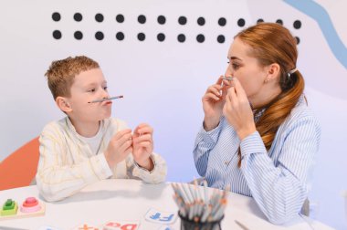 Genç bir çocuk, denge ve motor becerileri için üst dudaklarındaki kalemleri dengeleyen bir kadın profesyonelle konuşma terapisi egzersizleri öğreniyor.