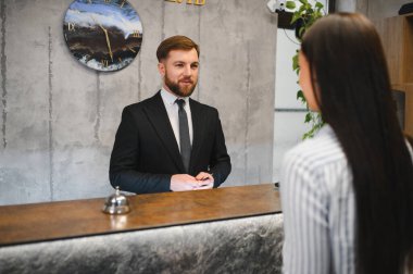 Profesyonel sakallı otel resepsiyonisti lobi masasında duruyor ve bir bayana konukseverlik hizmetinde yardım ediyor.