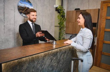 Dostane otel resepsiyonisti. Resepsiyonda gülümseyen bir kadına pano veriyor. Konukseverlik ve hizmet üzerine odaklanıyor.