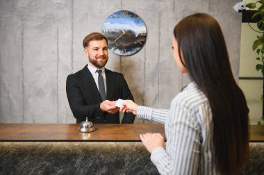 Gülümseyen erkek otel resepsiyonisti giriş yapan genç bir bayana yardım ediyor, resepsiyonda anahtar kartı veriyor.