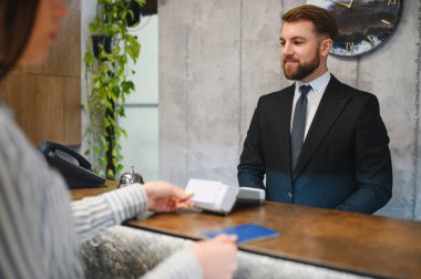 Otel resepsiyonunda çalışan takım elbiseli bir adam, oda kontrolünü tamamlayan kadına kart anahtarı veriyor.