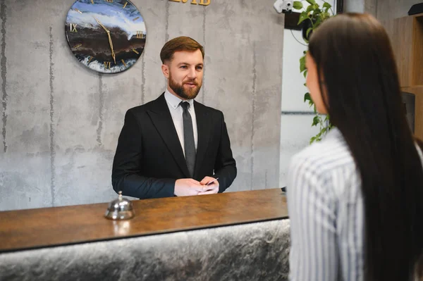Profesyonel sakallı otel resepsiyonisti lobi masasında duruyor ve bir bayana konukseverlik hizmetinde yardım ediyor.