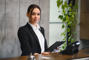 Resepsiyon masasının arkasında duran genç bir kadın elinde bir pano, gülümsüyor ve profesyonel konukseverlik hizmeti veriyor.