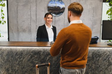 Modern bir resepsiyonda çalışırken gülümseyen otel resepsiyonisti konaklama için erkek bir konuğa yardım ediyor.