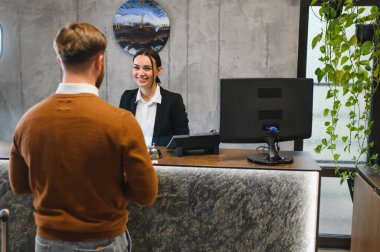 Otel resepsiyonunda durup gülümseyen kadın resepsiyonistle konuşan bir misafir. Hizmet, konukseverlik ve gezginlere yardım