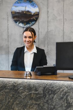 Modern bir otel resepsiyonunun arkasında duran gülümseyen kadın profesyonel konukseverlik hizmetleri sunuyor, müşterilere yardım etmeye hazır.