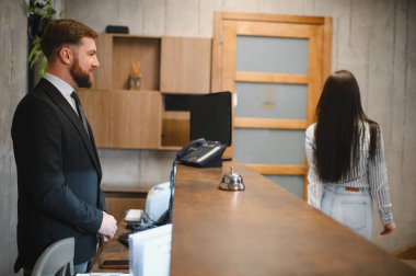Otel resepsiyonisti profesyonel hizmet sunuyor. Konukseverlik ortamında resepsiyondaki bir konukla konuşuyor.
