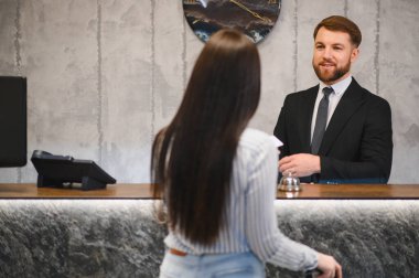 Dost canlısı, sakallı bir otel resepsiyonisti, modern resepsiyondaki bir konuğa yardımcı oluyor, kaliteli konukseverlik hizmeti veriyor.