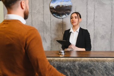 Konuk otel resepsiyonunda kadın resepsiyonistle görüşüyor, kontrol sırasında yardım alıyor. Konukseverlik hizmetine odaklan.