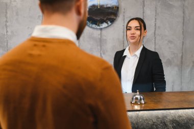 Bayan otel resepsiyonisti gülümsüyor, modern resepsiyonda erkek misafir ağırlıyor, kaliteli müşteri hizmeti sunuyor.