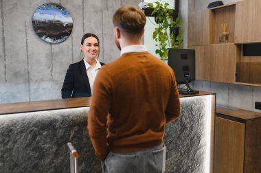 Çağdaş bir otel lobisine giriş sırasında erkek bir konuğa bavulla yardım ederken gülümseyen arkadaş canlısı bir bayan resepsiyonist.