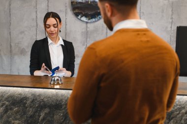 Resepsiyonist olarak çalışan genç bir kadın otel resepsiyonunda erkek misafirin pasaportunu kontrol ediyor, konukseverlik hizmeti veriyor.