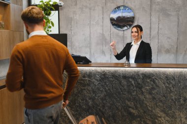 Otel resepsiyonunda duran, gülümseyen bir kadın yöneticiyle konuşan, kontrol için gelen bir misafir.