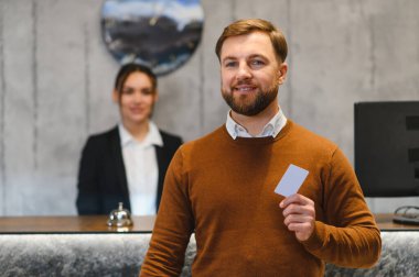 Gülümseyen bir adam, otel resepsiyonunda elinde boş bir anahtar kartıyla arka planda kadın resepsiyonist mükemmel bir hizmet sunuyor.