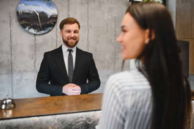 Gülümseyen profesyonel otel resepsiyonisti modern bir otel lobisinde genç bir bayanı selamlıyor ve ona yardım ediyor.