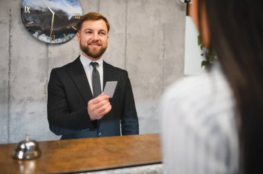 Gülümseyen erkek otel resepsiyonisti, danışmada bir konuğa yardım ediyor, kontrol sırasında anahtar kartı veriyor.