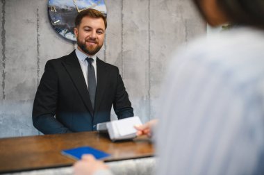 Otel resepsiyonisti dostça gülümserken konuğun resepsiyonda kontrolü tamamlamasına yardım ediyor ve servis sunuyor.