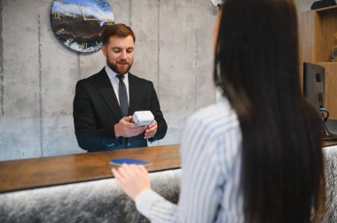 Otel resepsiyonisti bir müşteri için ödeme terminali tutuyor. Resepsiyonda düzgün bir kontrol işlemi yürütüyor.