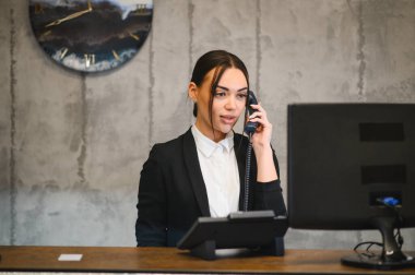 Genç bayan resepsiyonist, ofis tezgahında sabit bir telefonla konuşurken destek ve yardım teklif ediyor.