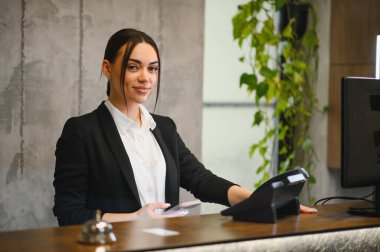 Otel resepsiyonunda çalışan genç bir kadın müşteri hizmetleri sunuyor ve ziyaretçi soruşturmalarını dostça bir gülümsemeyle yürütüyor.