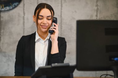 Resepsiyonda çalışan gülümseyen iş kadını ofis ortamında sabit telefon kullanarak müşteriyle iletişim kuruyor.