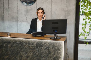 Gülümseyen kadın resepsiyonist modern bir otel resepsiyonunda müşteri hizmeti veriyor, elinde telefon ve yardımcı konuklar var.