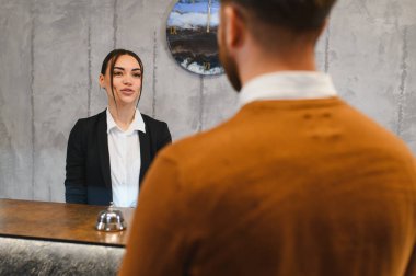Genç bayan resepsiyonist resepsiyonda erkek bir misafirle konuşuyor, konukseverlik ve bilgi kontrolü yapıyor.