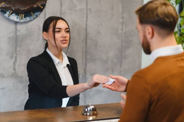 Kadın otel resepsiyonisti erkek bir müşteriye yardım ediyor. Resepsiyondaki kontrol sırasında giriş kartını teslim ediyor.