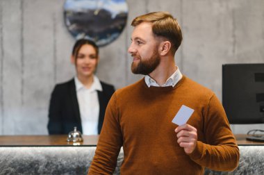 Konuk adamın elinde anahtar kartı var. Arka planda bir resepsiyonist ile kameraya bakıp gülümsüyor.