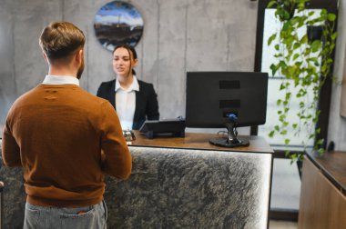 Otel girişini tamamlayan adam resepsiyonda. Dost canlısı bayan resepsiyonist müşteriye konukseverlik hizmeti veriyor.