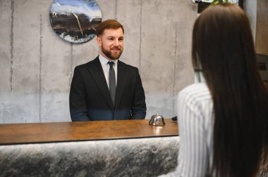 Gülümseyen erkek otel resepsiyonisti karşılama masasında duran bir müşteriye konukseverlik sunuyor.