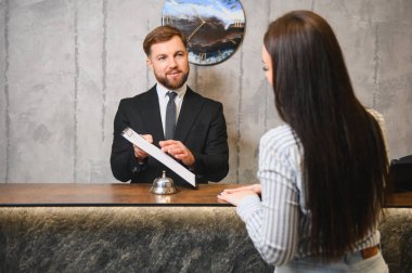 Profesyonel otel resepsiyonisti genç bir bayana resepsiyonda yardım ediyor. Konukseverlik ve müşteri hizmetlerine odaklanıyor.
