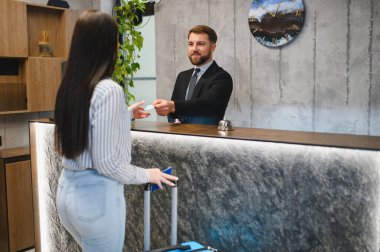 Modern otel resepsiyonuna gelen bayan misafir, yetişkin bir işadamı oda giriş kartı veriyor.