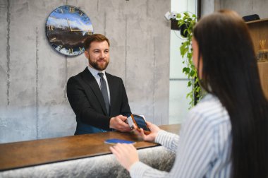 Gülümseyen otel resepsiyonisti müşteriye yardım ediyor akıllı telefon kullanıyor ve resepsiyondaki bağlantı kurulamıyor.